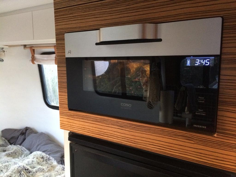 Inside an RV rental: a modern built-in microwave in a compact kitchen area with wood paneling, next to a cozy bed. — photo 12