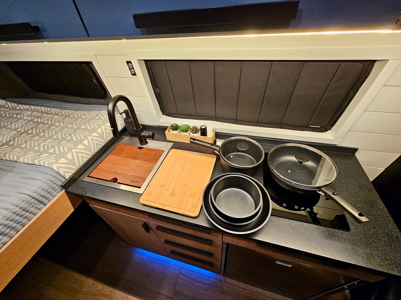 Compact RV rental kitchen and bedroom area featuring a sink, cooktop, bed, and stylish lighting for comfortable travel. — photo 17