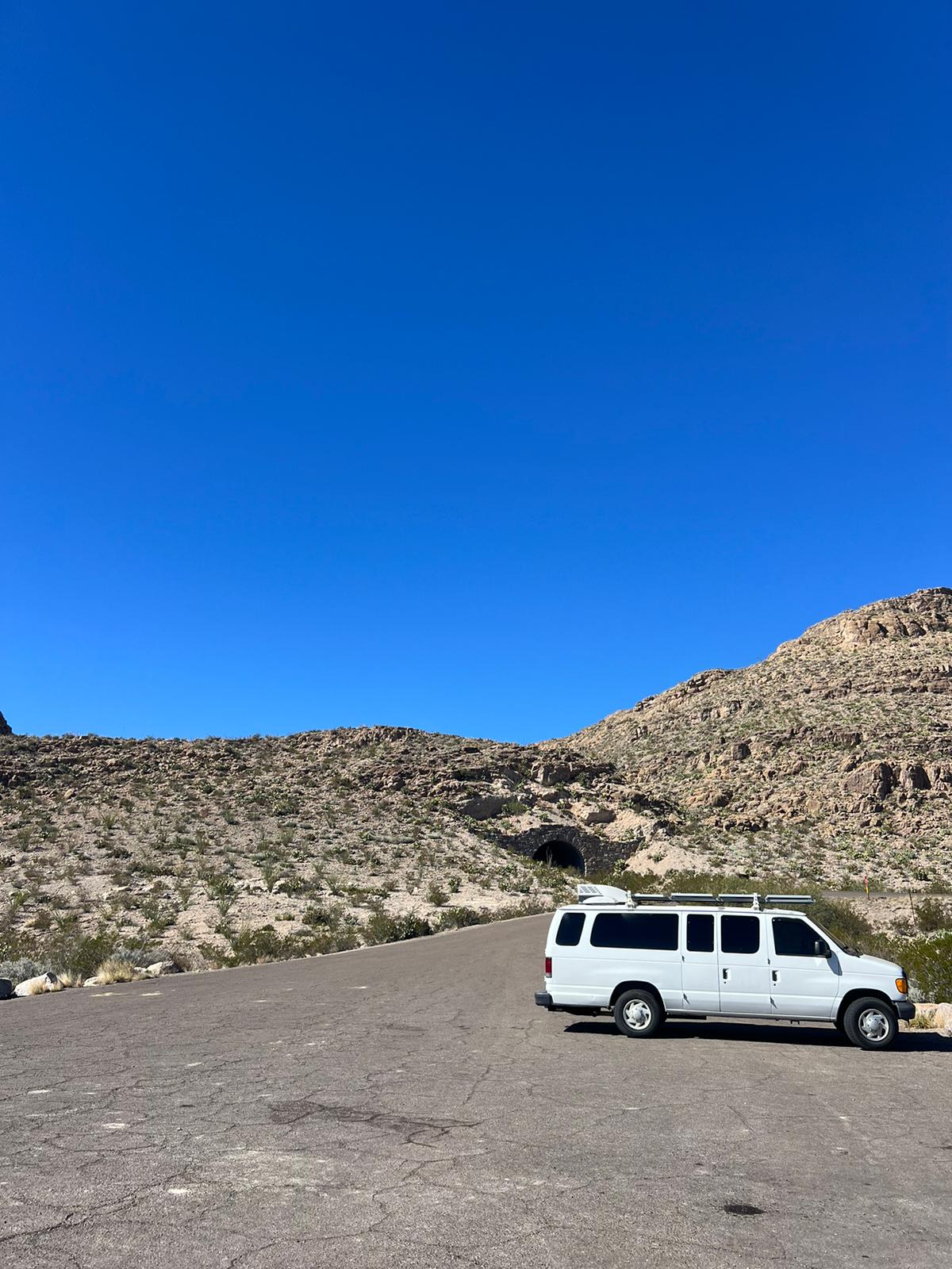 White RV rental van on a desert road with mountains & a tunnel under clear blue sky.