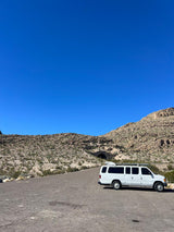 White RV rental van on a desert road with mountains & a tunnel under clear blue sky.