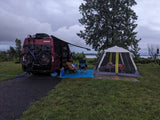 A cozy RV rental campsite with a red Winnebago van, bikes, chairs, and a screen tent under a cloudy sky.