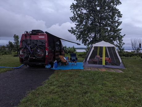 A cozy RV rental campsite with a red Winnebago van, bikes, chairs, and a screen tent under a cloudy sky.