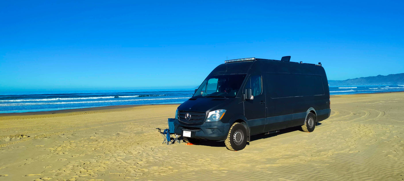 Dark RV rental Sprinter van parked on a sunny sandy beach next to the blue ocean. Camping chairs are out, ready for adventure.