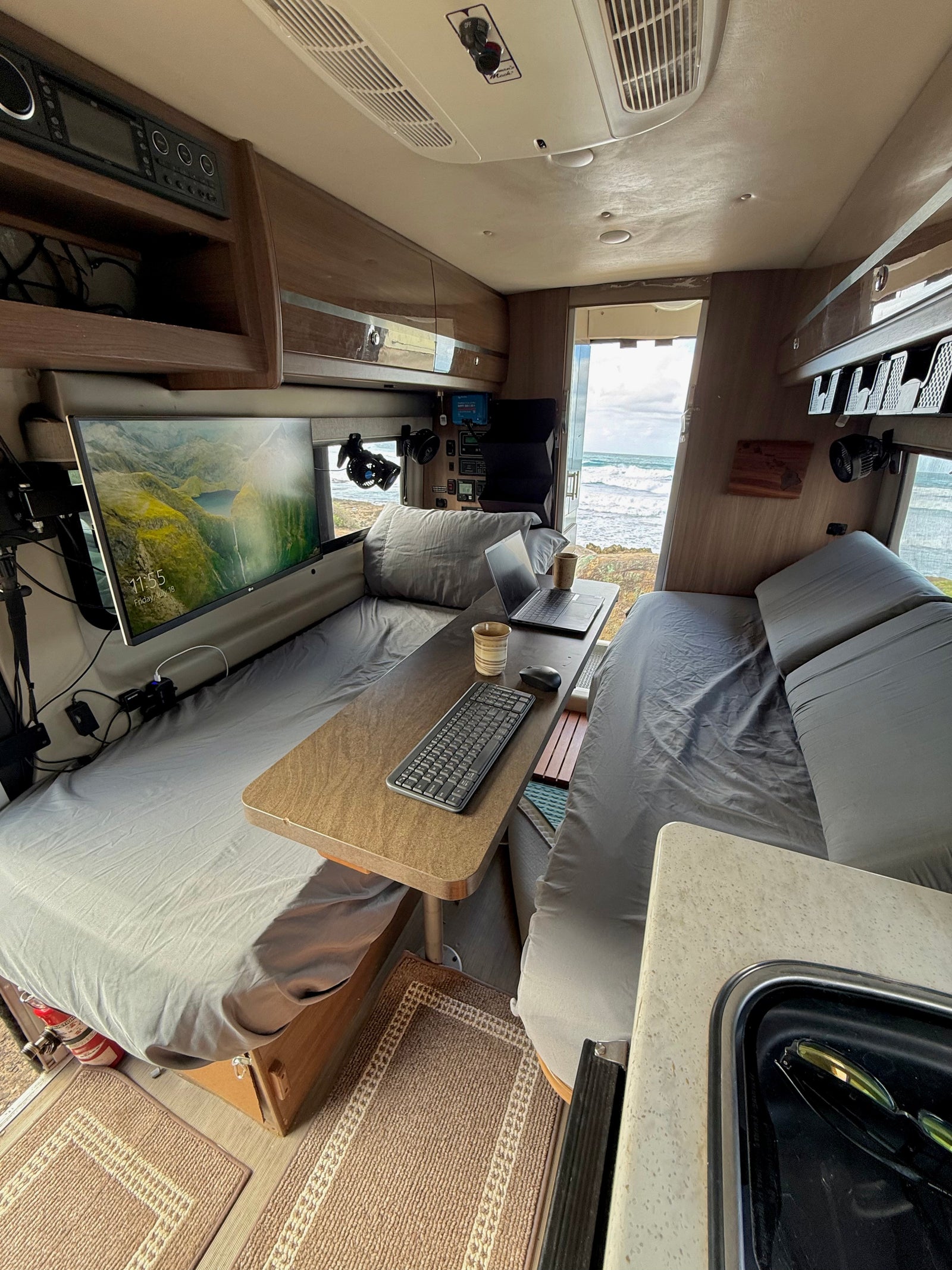 Cozy RV rental workspace with beds, a monitor, laptop, and scenic ocean views from the open rear.