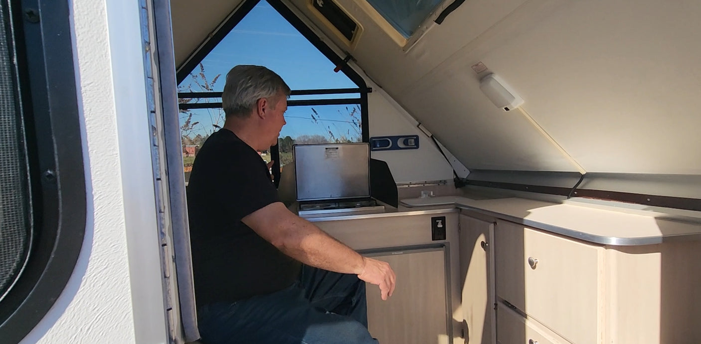A man is seated within an RV rental A-frame camper, with large windows overlooking trees. Features a compact kitchen & fridge.