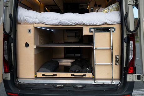 RV rental camper van's rear shows a raised bed, under-bed wooden storage with shelves, a pull-out drawer, and an access ladder.