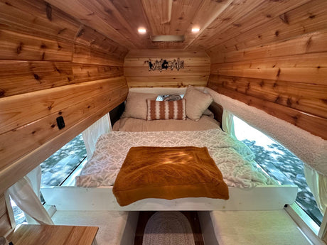 This RV rental features a cozy wood-paneled bedroom with a comfy bed & large windows, perfect for van life adventures.