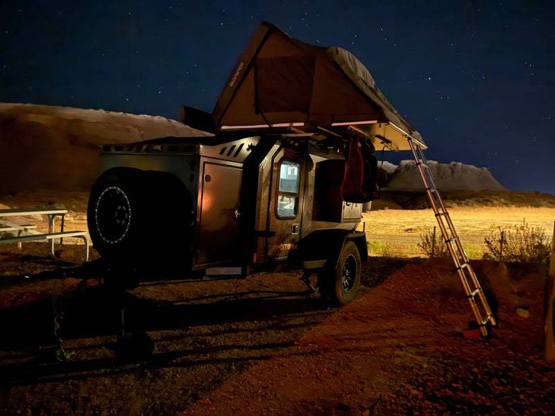  surrounded by vibrant golden and orange autumn trees under a cloudy sky.|Night camping with an RV rental overland trailer! Enjoy stargazing from this rooftop tent setup in a scenic desert landscape.|Black off-road rv rental "EXPEDITION 2.0" trailer with iKAMPER rooftop tent — photo 5