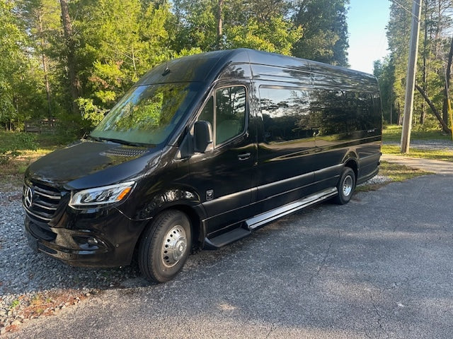 A sleek black Mercedes Sprinter van, ideal for rv rental, is parked on a gravel and asphalt patch by green trees. Perfect for travel.