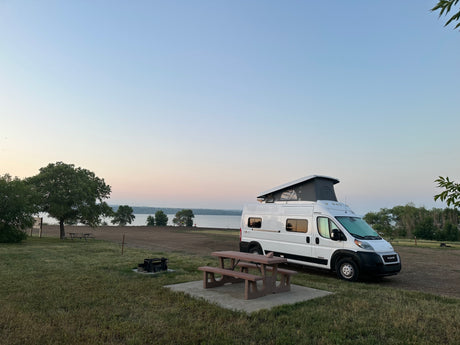 An rv rental camper van sits at a tranquil lakeside campsite with a picnic table and fire pit.