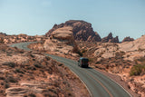 Black RV rental drives on a scenic desert road with red rock formations under a clear sky.