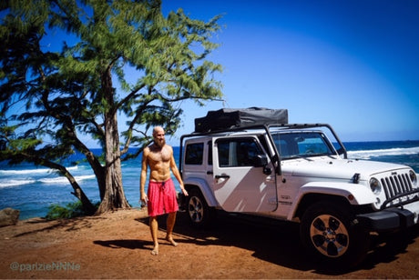Man by white Jeep Wrangler with rooftop tent on a sunny coast. Consider this style of RV rental for adventure.