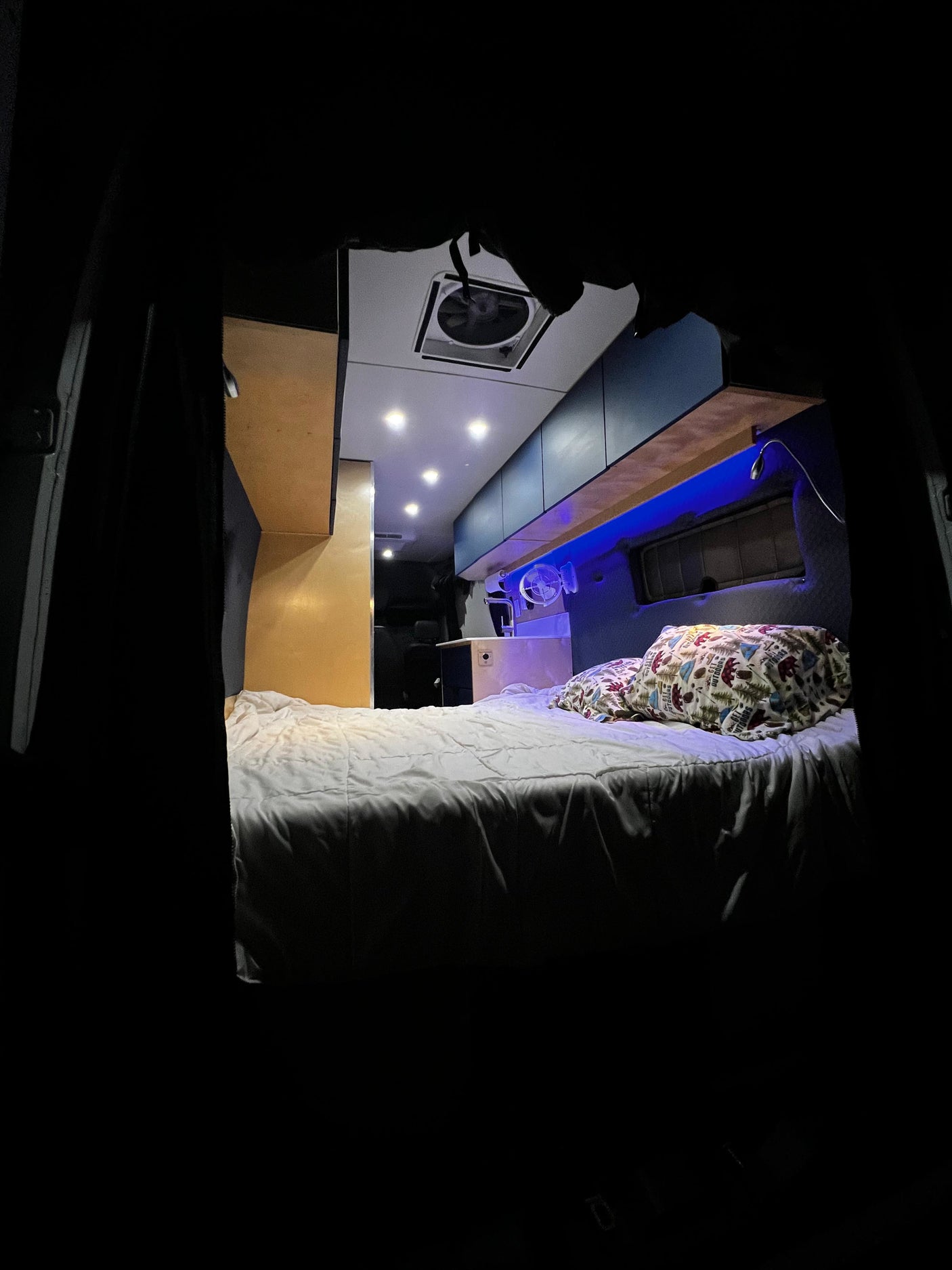 Inside this rv rental, a cozy bed with patterned pillows, blue accent lighting, overhead spotlights, and a ceiling fan create comfort.