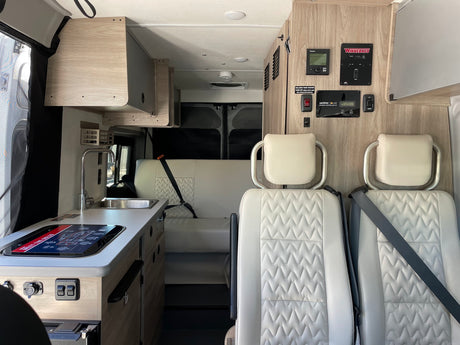 Inside a modern RV rental: light wood cabinets, white quilted seats, kitchen, and system controls.