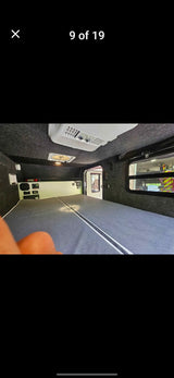 Inside this RV rental: a spacious gray bed, dark upholstered interior with AC, fan, controls, and a window view.