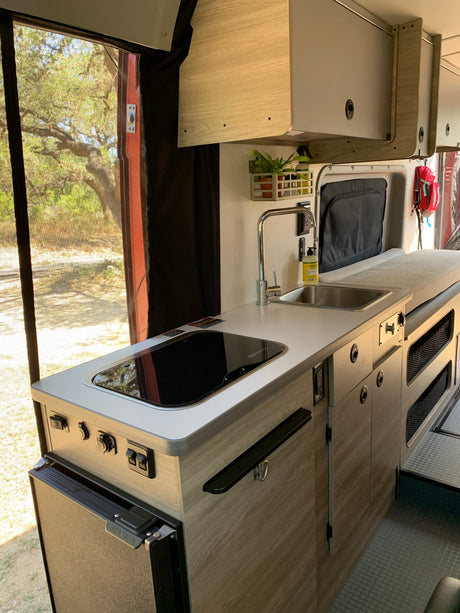 Modern RV rental interior with a compact kitchen (sink, cooktop, fridge) and comfortable bed, overlooking nature.