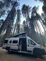 RV rental van parked in a serene tall pine forest with a mountain vista. Pop-top raised for camping.
