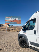 White RV rental van parked at the iconic "Welcome to Utah" sign, showcasing desert scenery under a clear blue sky.