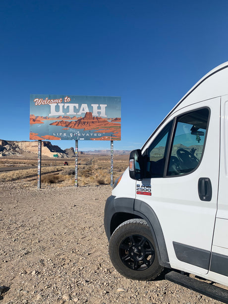 White RV rental van parked at the iconic "Welcome to Utah" sign, showcasing desert scenery under a clear blue sky.