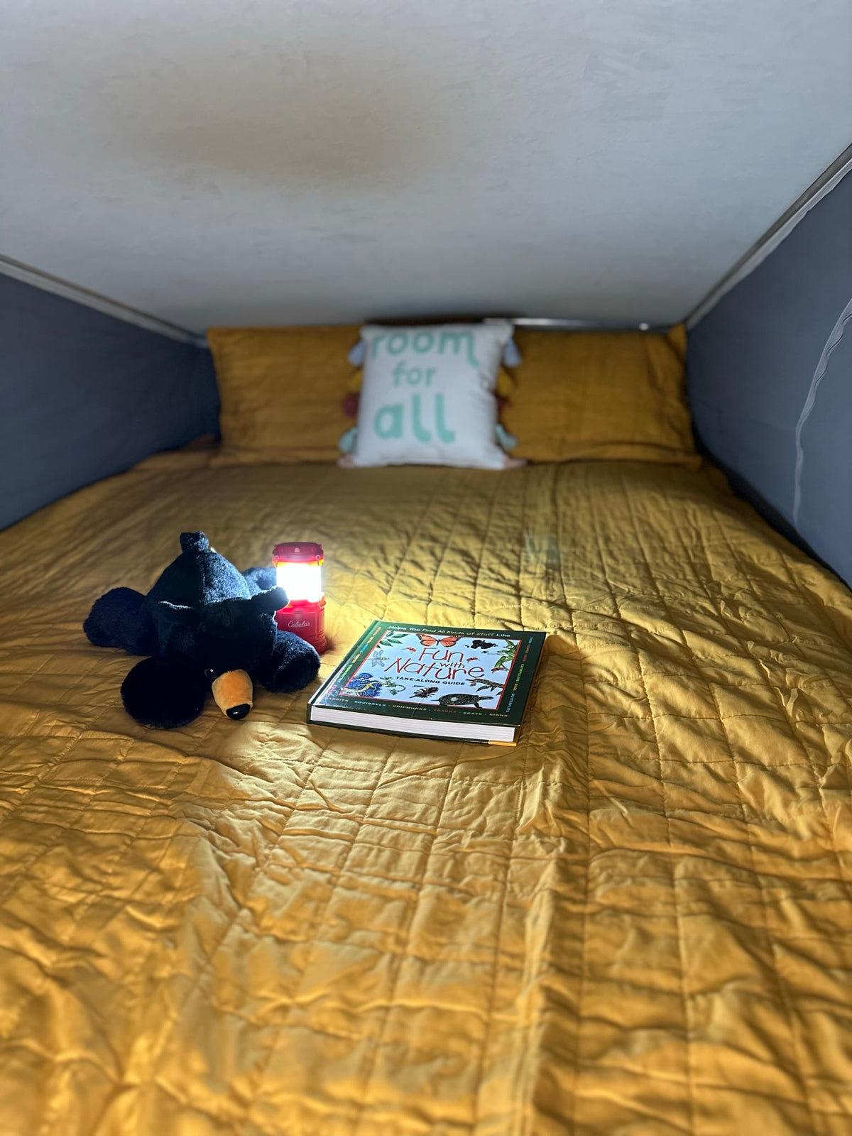 Cozy RV rental bed with yellow quilt, plush bear, lit lantern, and a 'Fun with Nature' book. Perfect for reading on camping adventures. — photo 12
