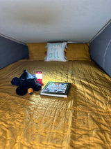 Cozy RV rental bed with yellow quilt, plush bear, lit lantern, and a 'Fun with Nature' book. Perfect for reading on camping adventures.