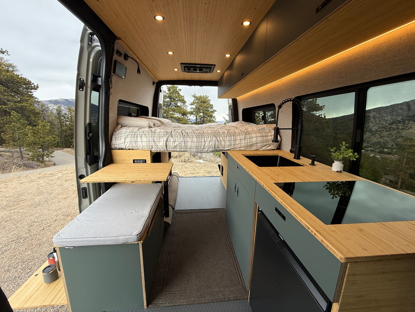 This RV rental interior shows a cozy bed, modern kitchen with bamboo counters, and scenic mountain views from open doors.