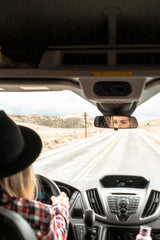 Driving an RV rental, a person in a hat is seen from behind the wheel, with eyes reflected in the rearview mirror on a scenic road.