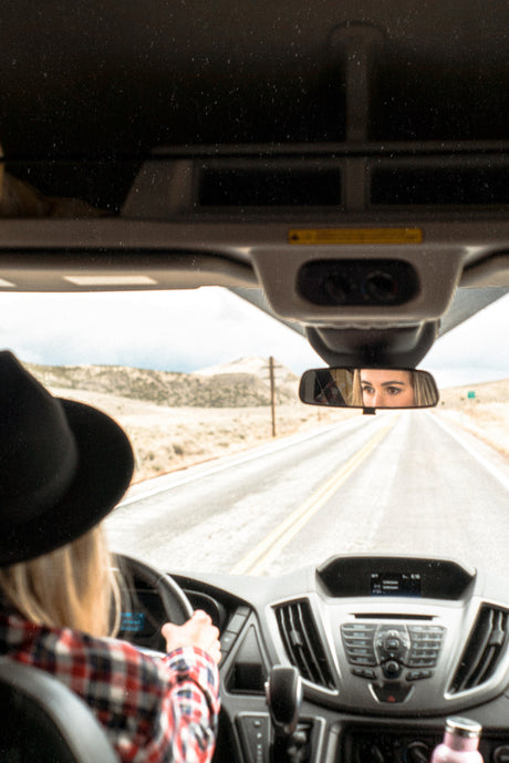 Driving an RV rental, a person in a hat is seen from behind the wheel, with eyes reflected in the rearview mirror on a scenic road.
