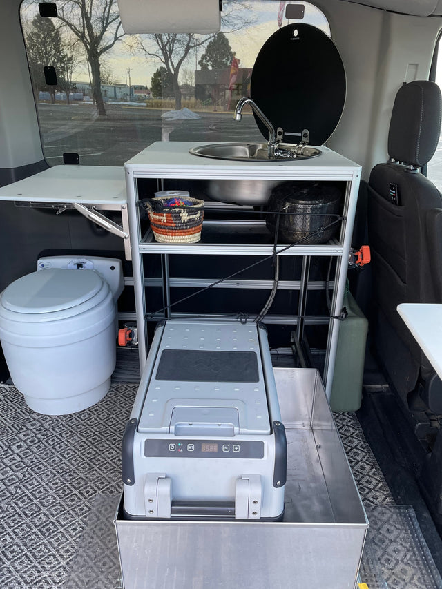 Compact RV rental van interior with a portable toilet, kitchen sink, cooler, and folding table for travel.