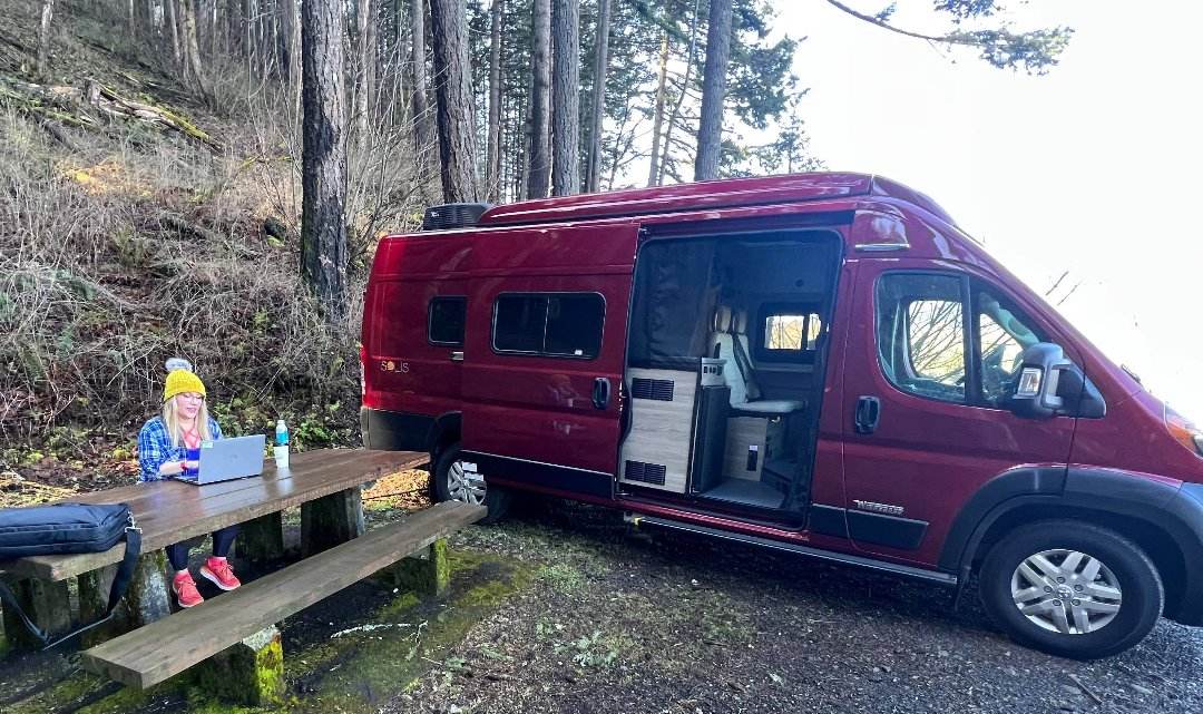  parked by a lake amidst dry brush under a cloudy sky. An RV rental opportunity.|Woman remote working at a picnic table beside her red Solis camper van. Ideal for an RV rental adventure!|Red Winnebago Solis RV rental with raised pop-top by a scenic river. Ready for outdoor adventure under cloudy skies.|A red RV rental van parked on a winding road next to a lake — photo 2