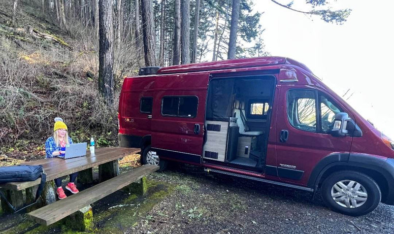  parked by a lake amidst dry brush under a cloudy sky. An RV rental opportunity.|Woman remote working at a picnic table beside her red Solis camper van. Ideal for an RV rental adventure!|Red Winnebago Solis RV rental with raised pop-top by a scenic river. Ready for outdoor adventure under cloudy skies.|A red RV rental van parked on a winding road next to a lake — photo 2