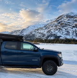 This blue truck camper setup, ideal for an RV rental, is parked in a snowy mountain landscape by a frozen lake.