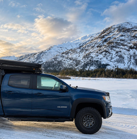This blue truck camper setup, ideal for an RV rental, is parked in a snowy mountain landscape by a frozen lake.