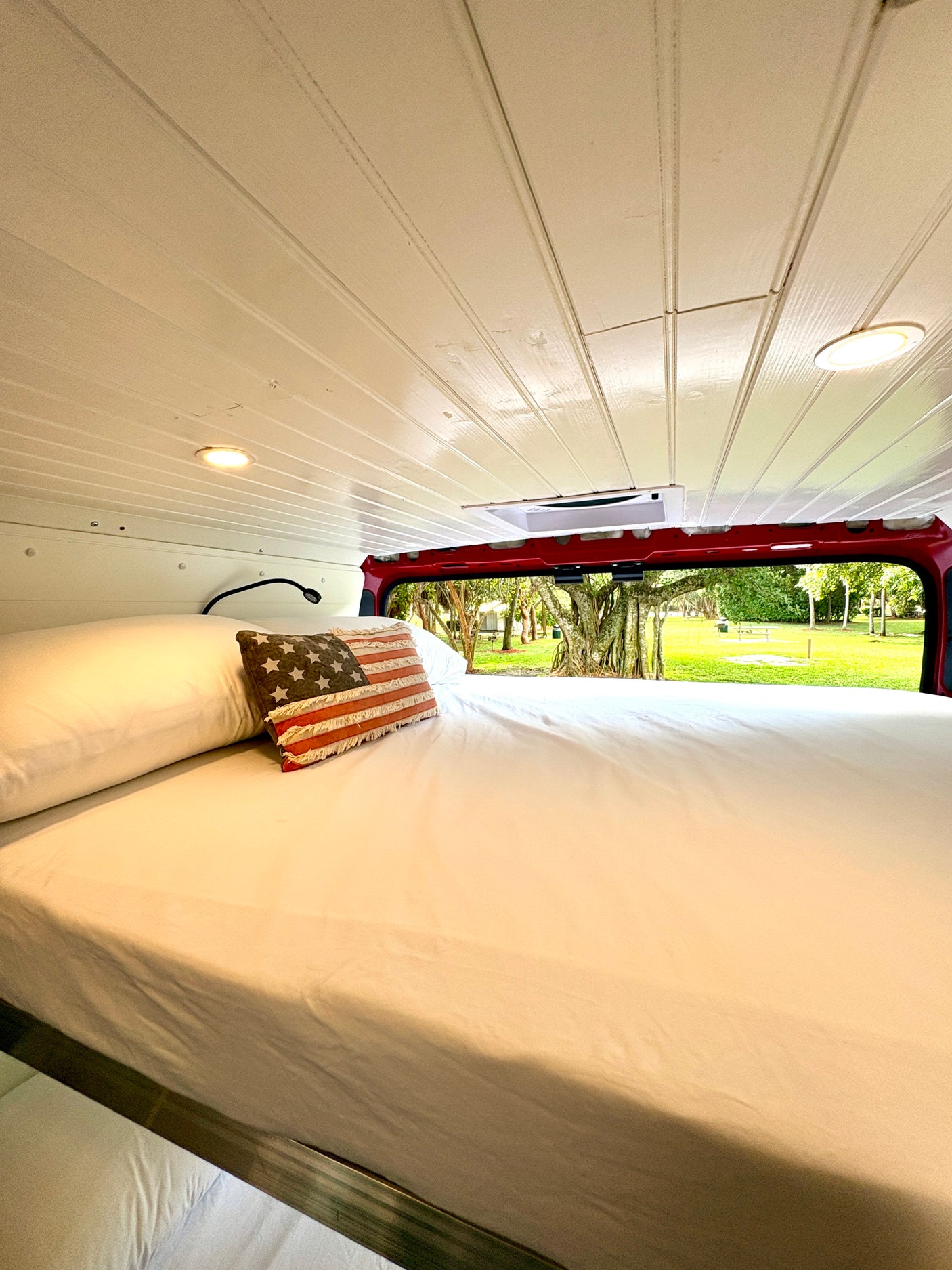 Cozy RV rental bedroom with white sheets, patriotic pillow, and a peaceful park view out the back.
