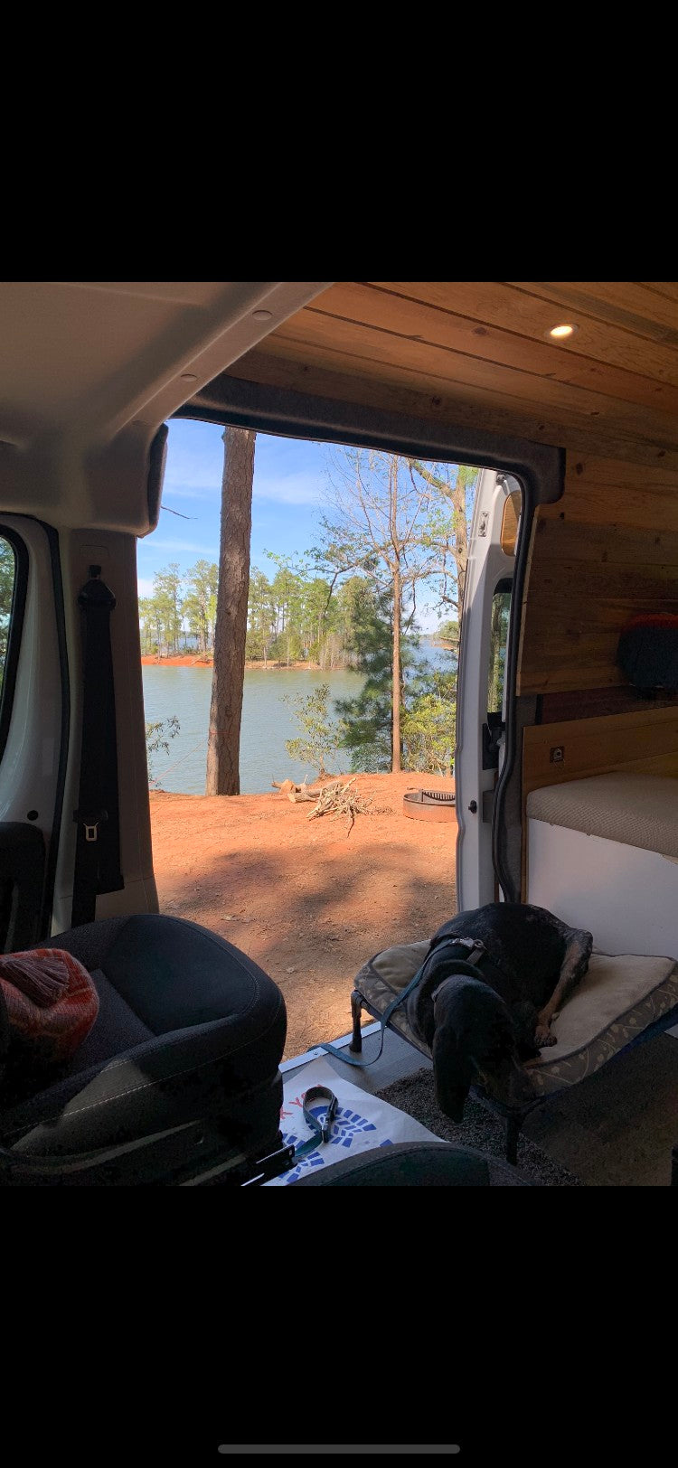 Scenic rv rental view: tranquil lake, forest, and a dog napping by the open van doors. Ideal for nature lovers.