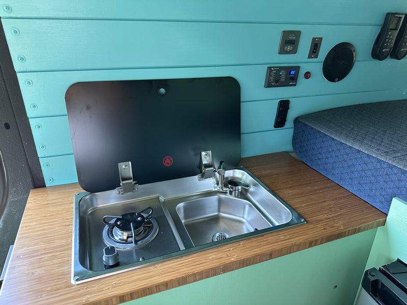 RV rental interior featuring a teal kitchenette with stove and sink, plus essential power controls and a bed. — photo 9