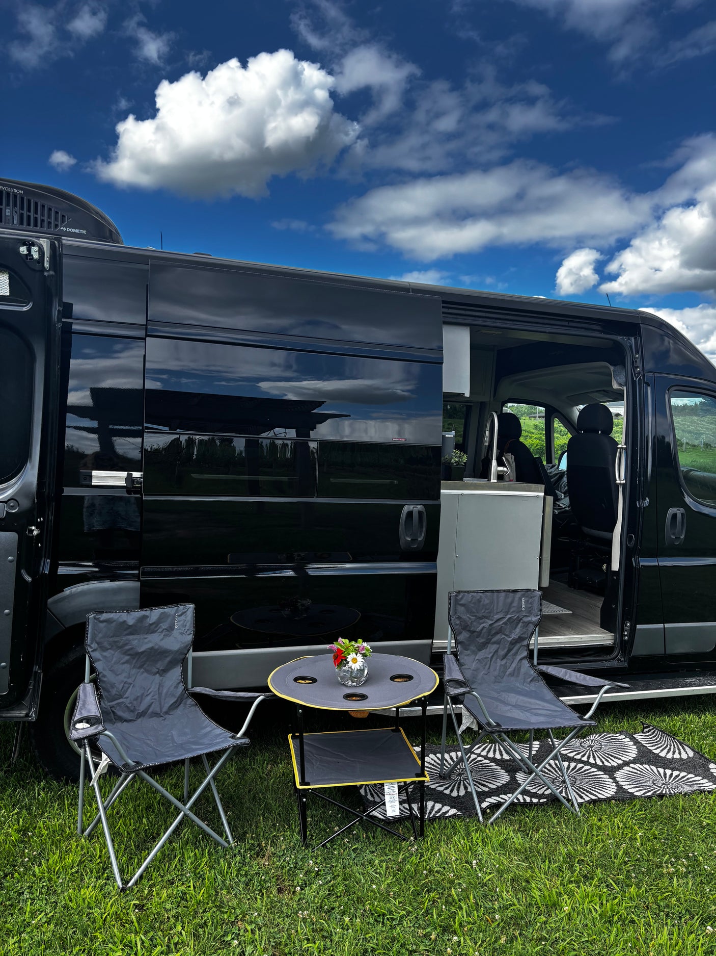 Black rv rental camper van with outdoor seating on green grass under a blue sky, perfect for a sunny day getaway.