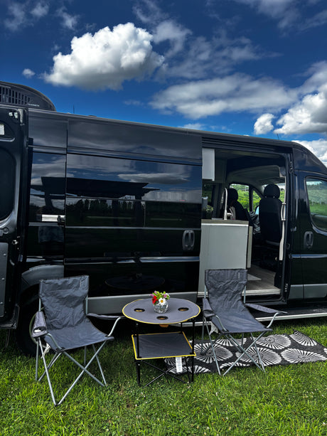 Black rv rental camper van with outdoor seating on green grass under a blue sky, perfect for a sunny day getaway.