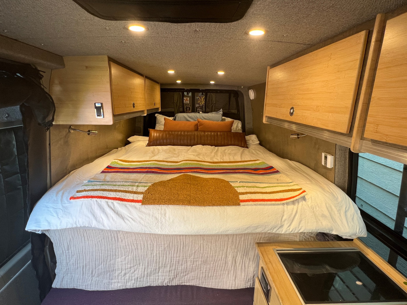 A bright, organized RV rental interior features a comfortable bed, wooden overhead cabinets, and integrated cooktop. — photo 12