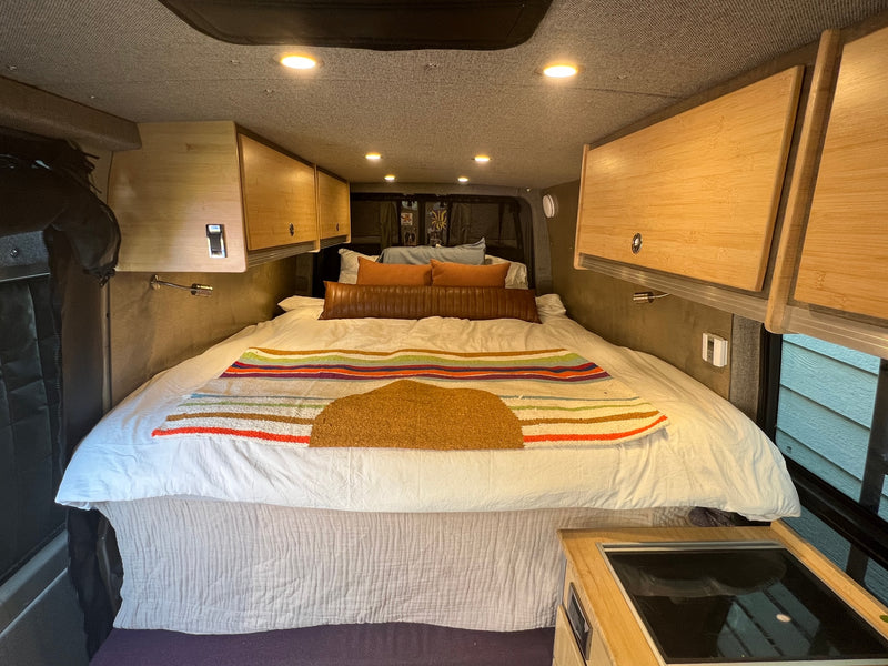 A bright, organized RV rental interior features a comfortable bed, wooden overhead cabinets, and integrated cooktop. — photo 12