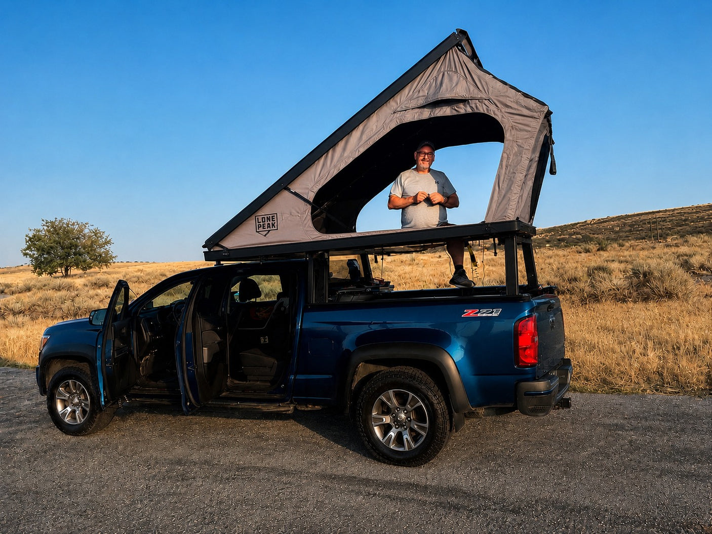 Man in a Lone Peak rooftop tent on a blue pickup truck, ready for an overland adventure. Consider this RV rental option!