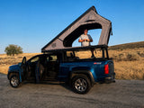Man in a Lone Peak rooftop tent on a blue pickup truck, ready for an overland adventure. Consider this RV rental option!