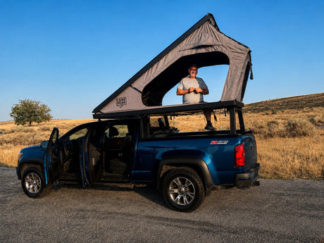 Man in a Lone Peak rooftop tent on a blue pickup truck, ready for an overland adventure. Consider this RV rental option!