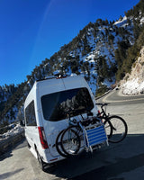 White RV rental van, bikes, and chair on rack, parked on a snowy mountain road under a clear sky.