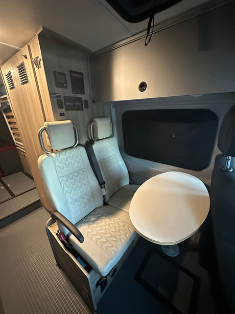 Modern RV rental interior features two light-colored seats, a round table, and wall-mounted control panels.