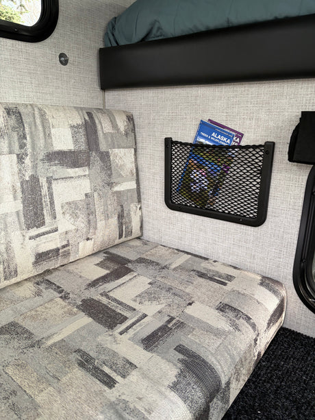 Cozy RV rental interior with patterned bench, wall storage net holding Alaska books, and an upper bunk for travel.