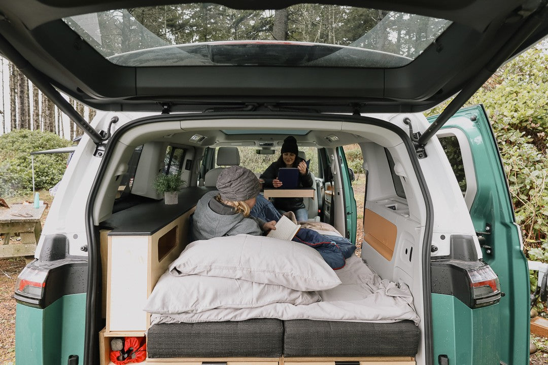 RV rental: From the open tailgate, two people relax inside a camper van; one reads, one uses a tablet.
