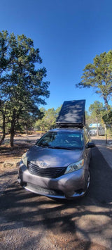 Grey minivan with rooftop tent pitched at a sunny campground surrounded by trees. A great RV rental alternative for outdoor adventures.