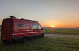 An rv rental, a red camper van, sits peacefully in a grassy field, bathed in the warm glow of sunrise over crops.