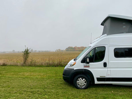 This white Winnebago RV rental, with its pop-top open, is set on green grass, overlooking a field under an overcast sky.